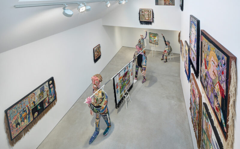 Art gallery interior featuring colorful textile artworks on walls and sculptural figures interacting with the space.