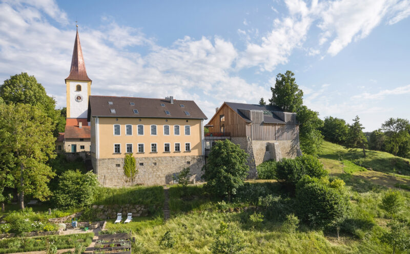 Aerial view of a building complex with a church tower, surrounded by greenery and gardens.
