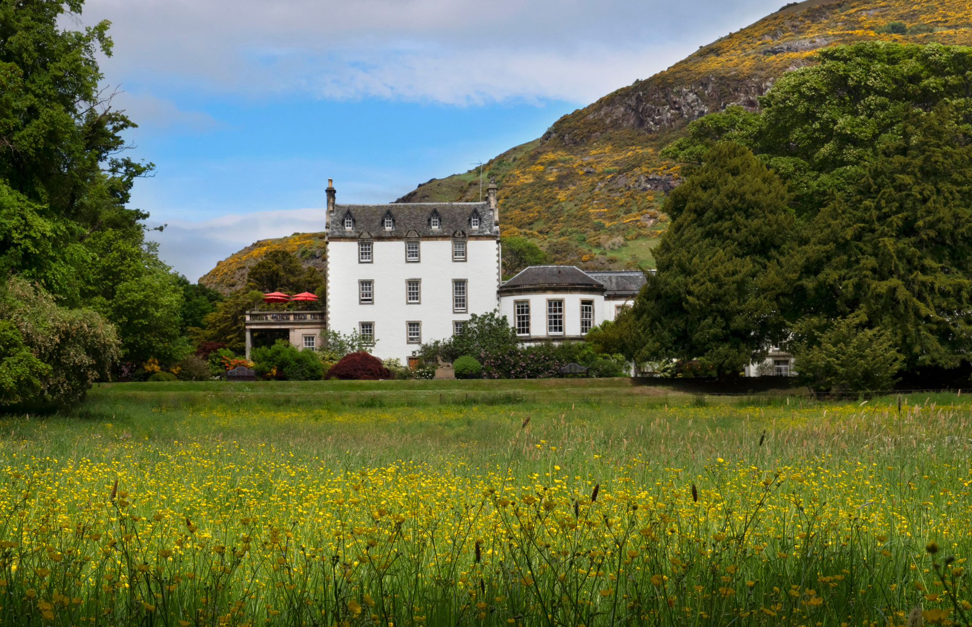 Prestonfield House surrounded by lush greenery and wildflowers, with hills in the background under a partly cloudy sky.
