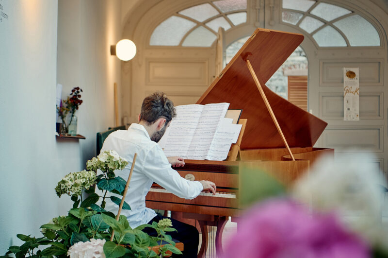 A musician playing a grand piano in a well-lit room, surrounded by flowers and sheet music.