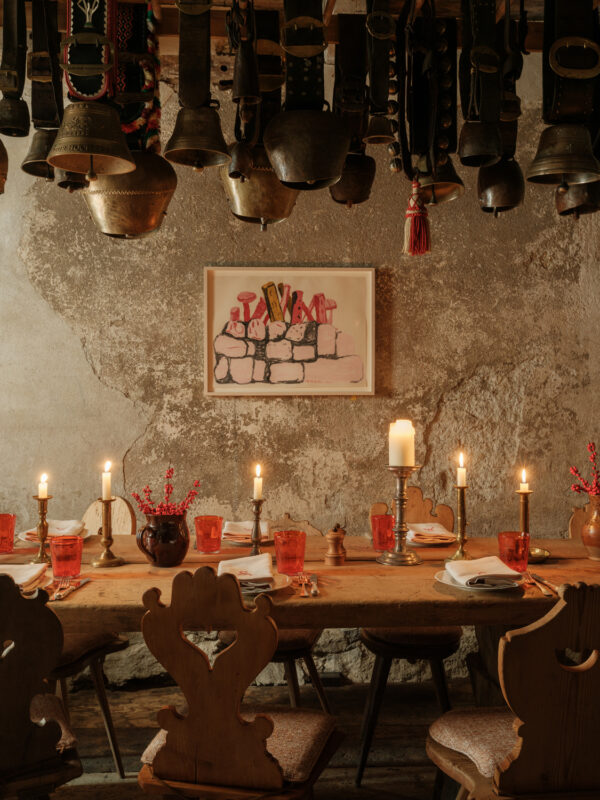 Rustic dining table set with red glasses and candles, adorned with a wall art piece and hanging bells in a cozy restaurant.