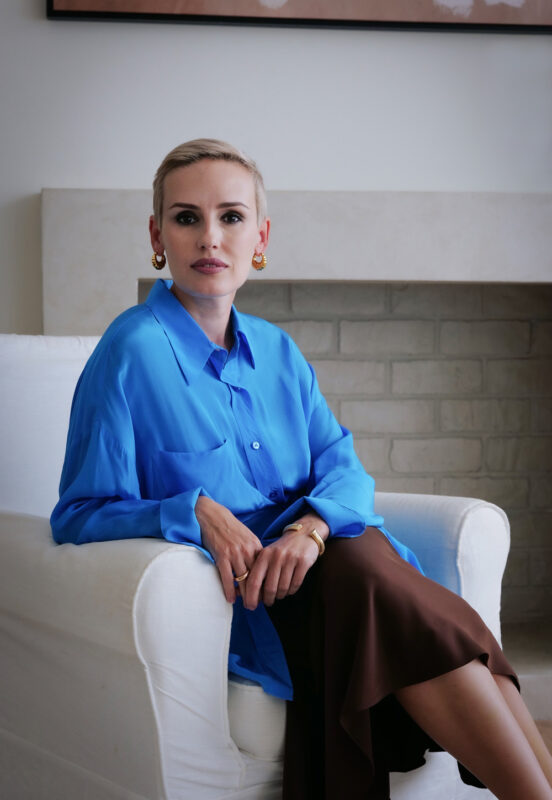 Woman with short blonde hair wearing a blue blouse and brown skirt, seated on a white chair in a bright room.
