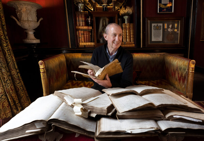 Man seated on a couch in Prestonfield House, holding open a book, surrounded by stacks of historical manuscripts.