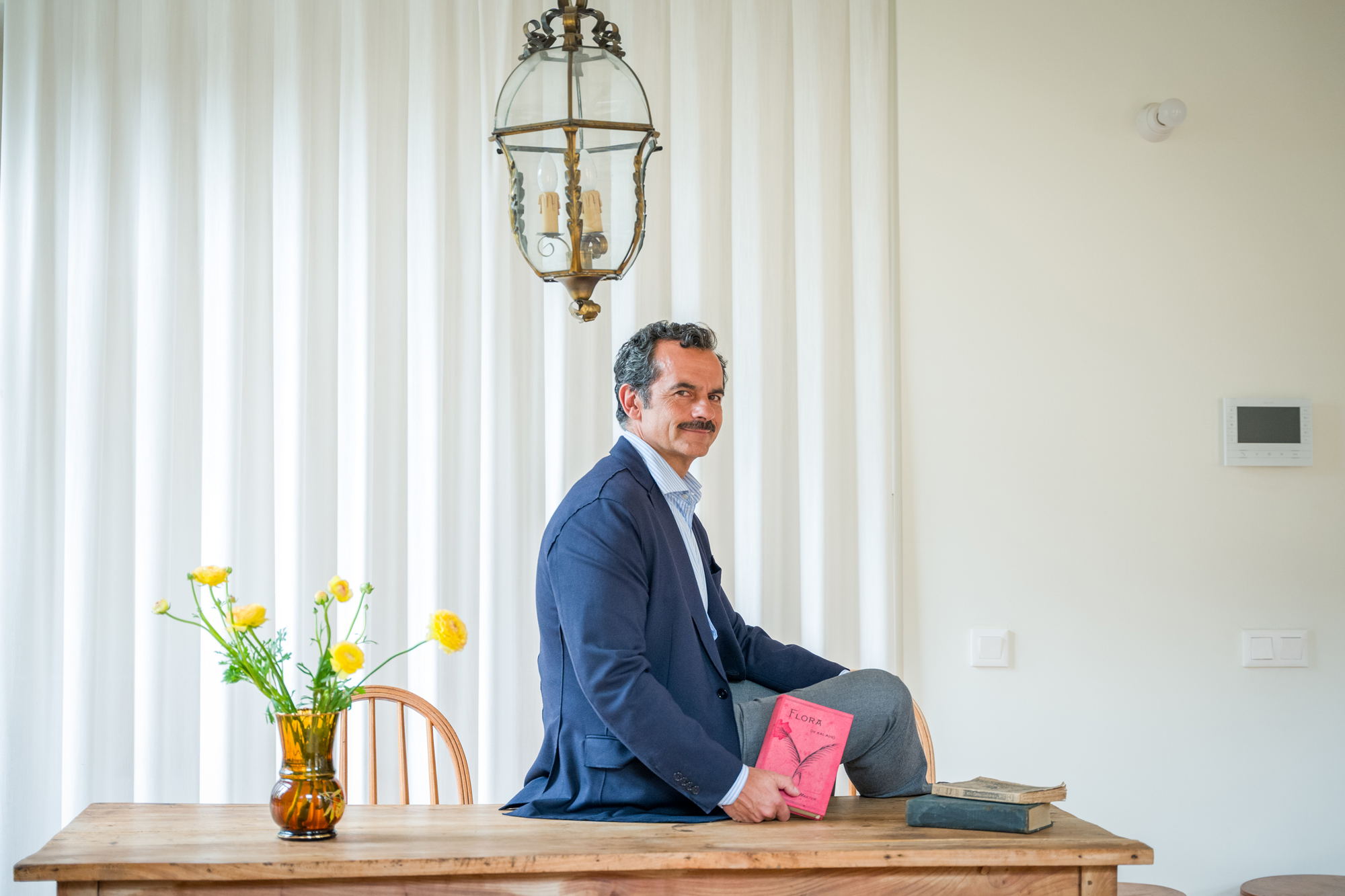 Man sitting on a wooden table, holding a pink book, with a vase of yellow flowers and a pendant light in a bright room.