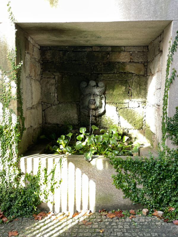 Stone wall alcove with a decorative water fountain featuring a face, surrounded by greenery and sunlight.