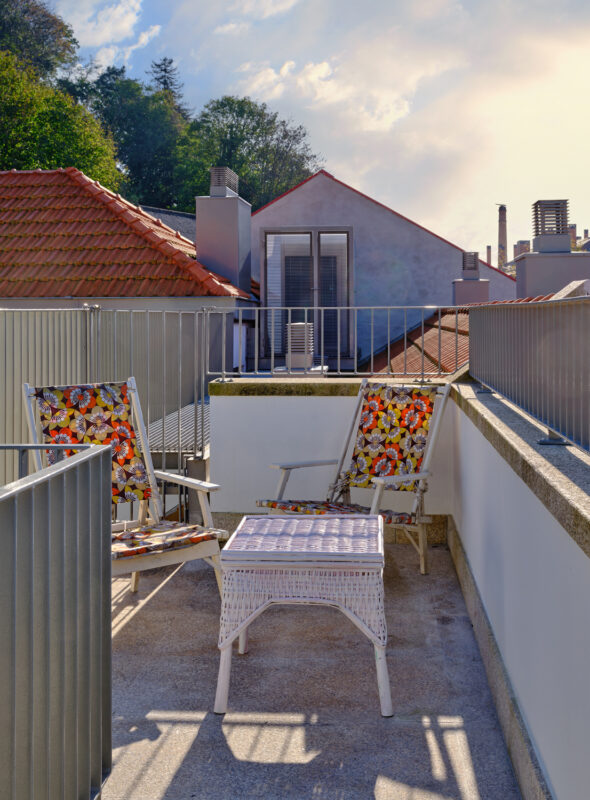 Rooftop terrace with two floral-patterned chairs and a small table, overlooking rooftops and trees under a blue sky.