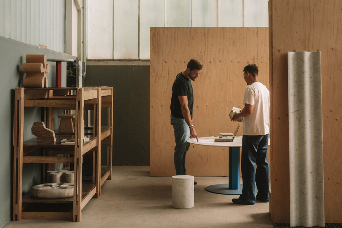 Two men examine materials on a round table in a workshop, surrounded by wooden shelves and marble samples.