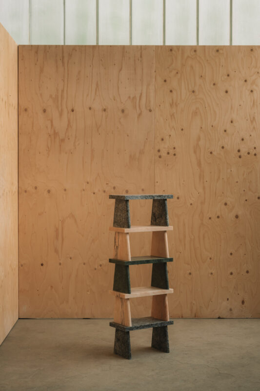 A wooden shelf with four tiers, featuring a mix of light wood and dark stone-like materials, set against a plywood wall.