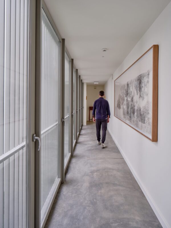 Long corridor with large windows, a person walking away, and a framed artwork on the wall.