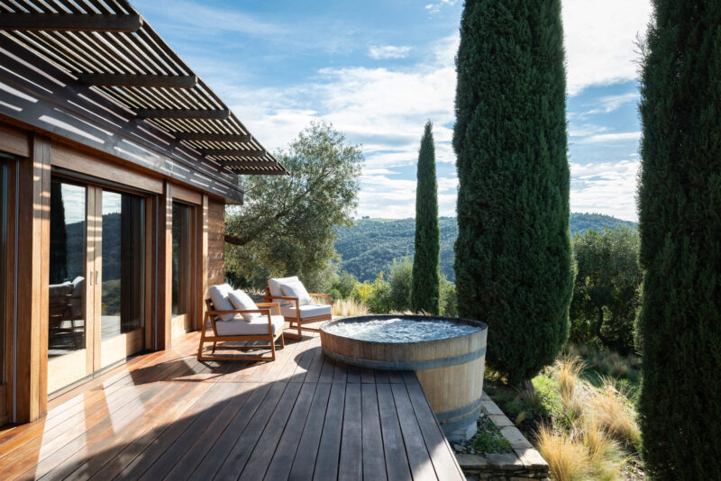 Wooden deck with lounge chairs and a hot tub, surrounded by cypress trees and hills under a blue sky.