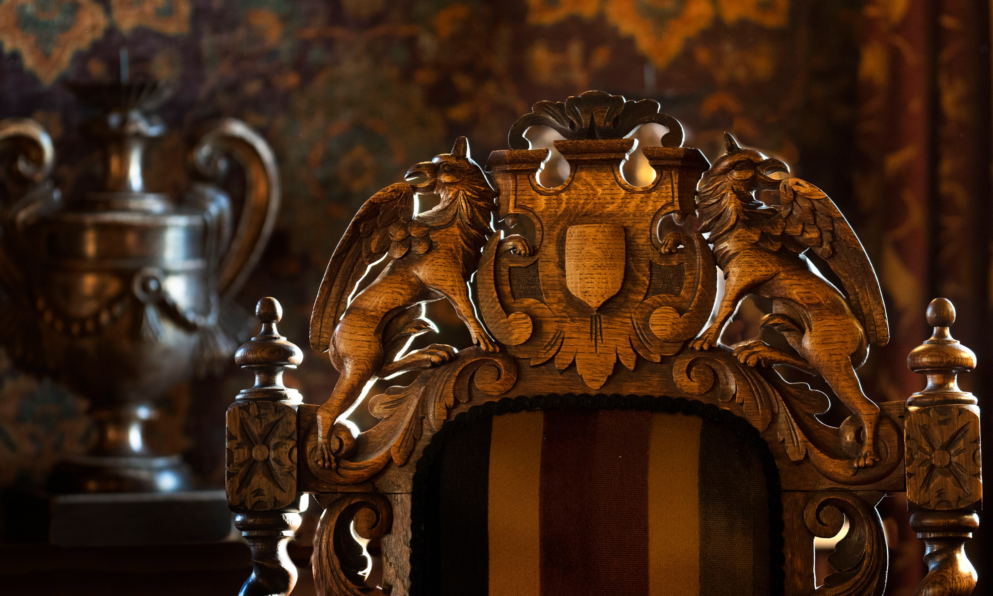 Intricately carved wooden chair with griffin motifs, set against a richly patterned backdrop and a decorative urn.