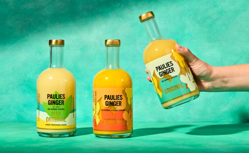 Three bottles of Paulies Ginger drink on a teal background, with one bottle being held and labeled "Original.