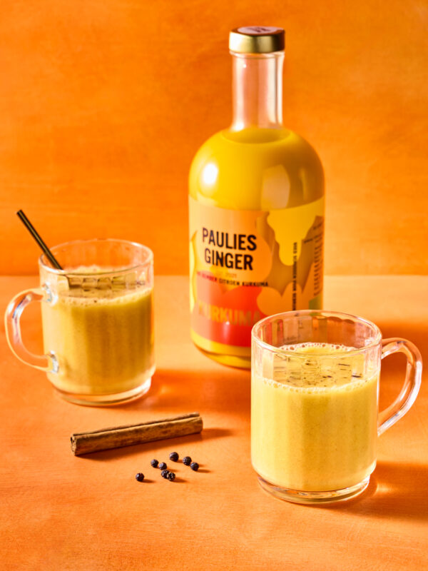 Two clear mugs filled with a creamy ginger drink, accompanied by a bottle labeled "Paulies Ginger" and cinnamon sticks on ...