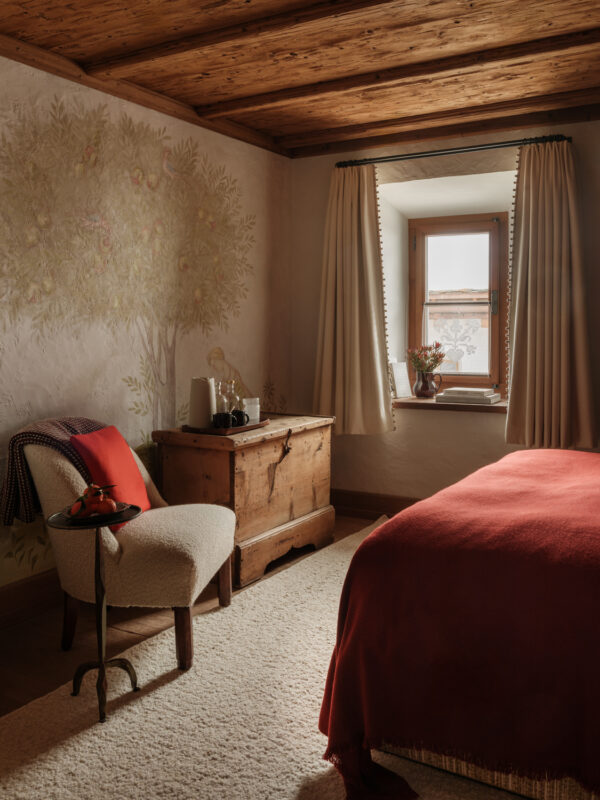 Cozy bedroom featuring a wooden bed with a red blanket, a vintage chest, and a chair, adorned with pomegranate-themed decor.