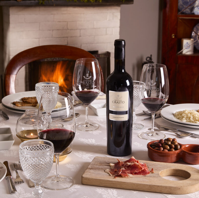 Table set with wine glasses, a bottle of Quinta do Crasto wine, cured meat, olives, and a cozy fireplace in the background.