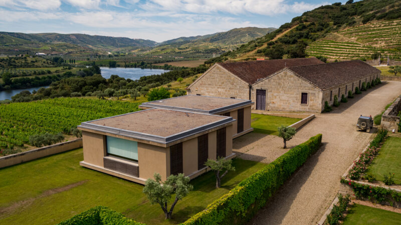 Modern winery buildings surrounded by lush vineyards and rolling hills, with a river in the background at Quinta do Vale M...