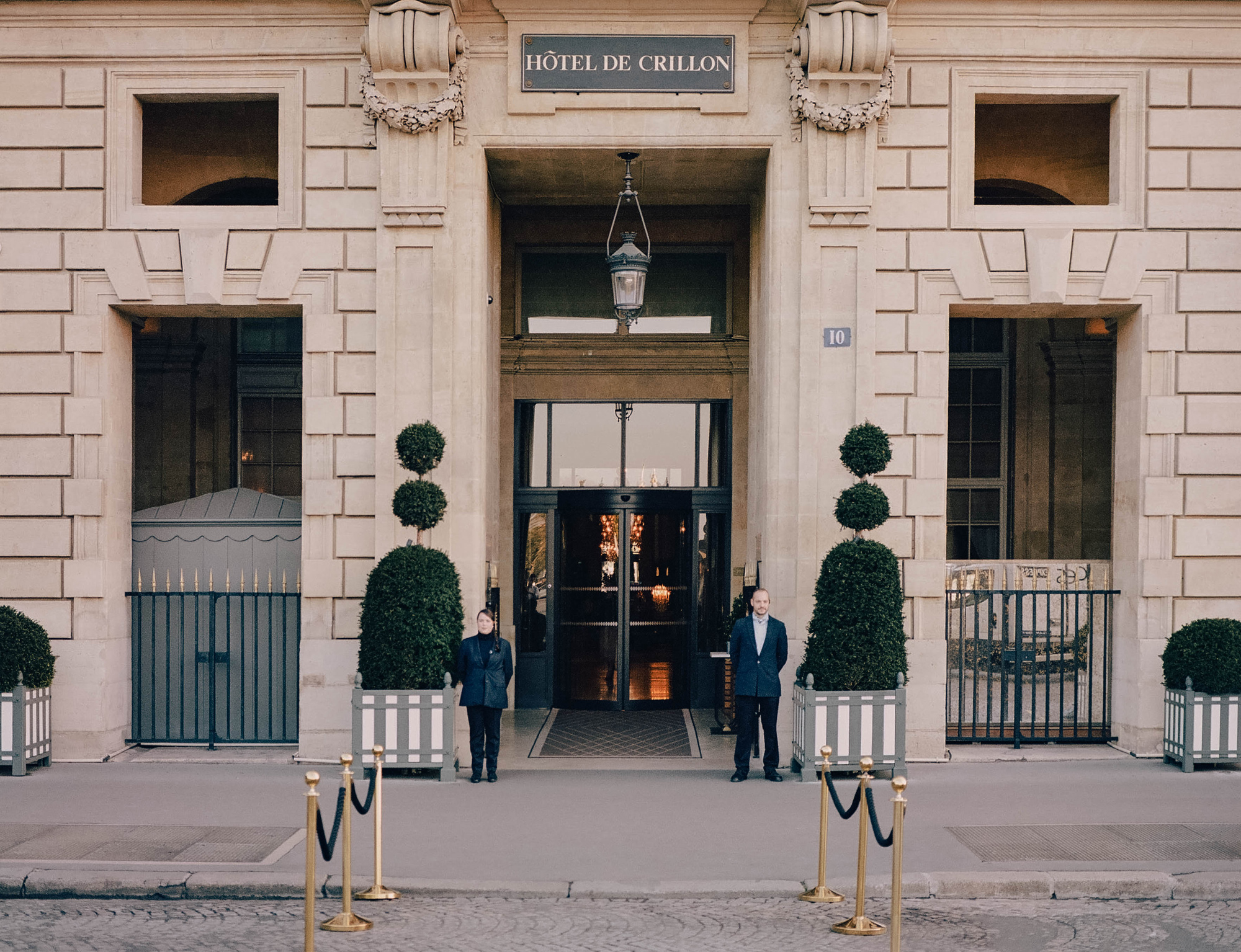 Facade of Hôtel de Crillon with two staff members standing by entrance, flanked by neatly trimmed topiary.