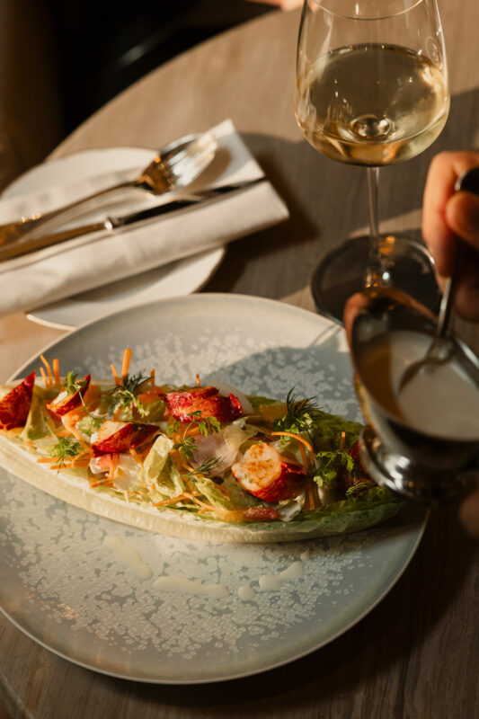 Lobster salad served on a white plate, accompanied by a glass of white wine and cutlery on a wooden table.