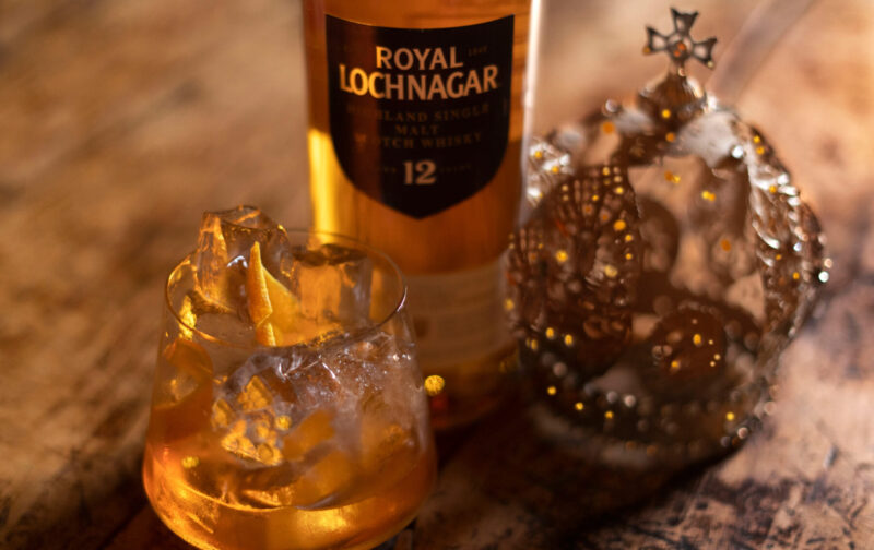Cocktail glass with ice beside a bottle of Royal Lochnagar whisky and a decorative crown on a wooden surface.