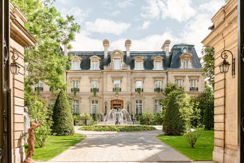 Elegant mansion with a central fountain, framed by manicured gardens and trees, viewed through ornate gates.