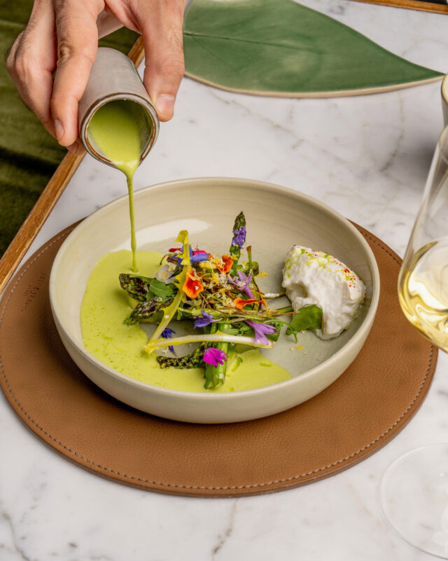 Asparagus dish with edible flowers, drizzled with green sauce, served with a dollop of cream on a marble table.