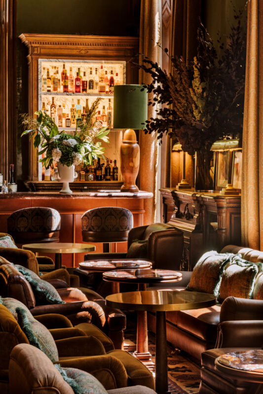 Elegant bar interior at Saint James Paris, featuring plush seating, a well-stocked backlit bar, and decorative floral arra...