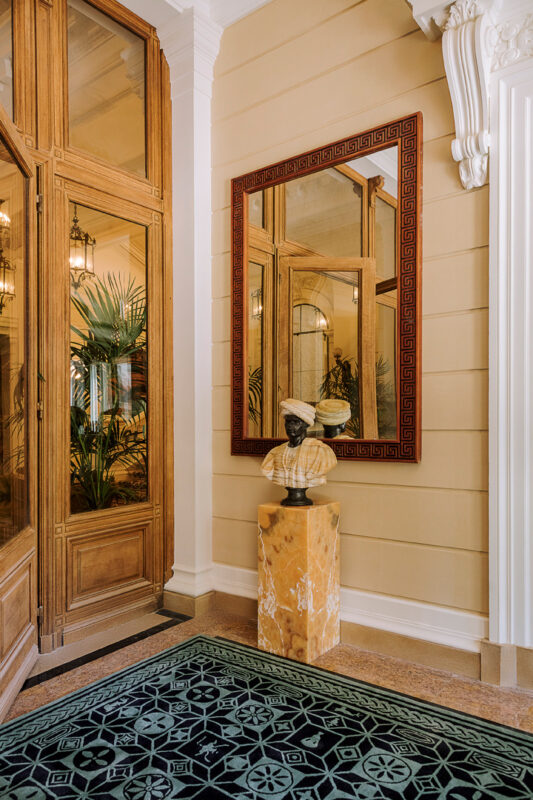 Elegant entrance of Saint James Paris featuring a large mirror, a bust on a marble pedestal, and decorative plants.