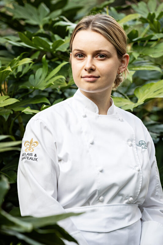 Pastry chef Coline Doussin in a white chef's coat, standing among lush green foliage at Saint James Paris.