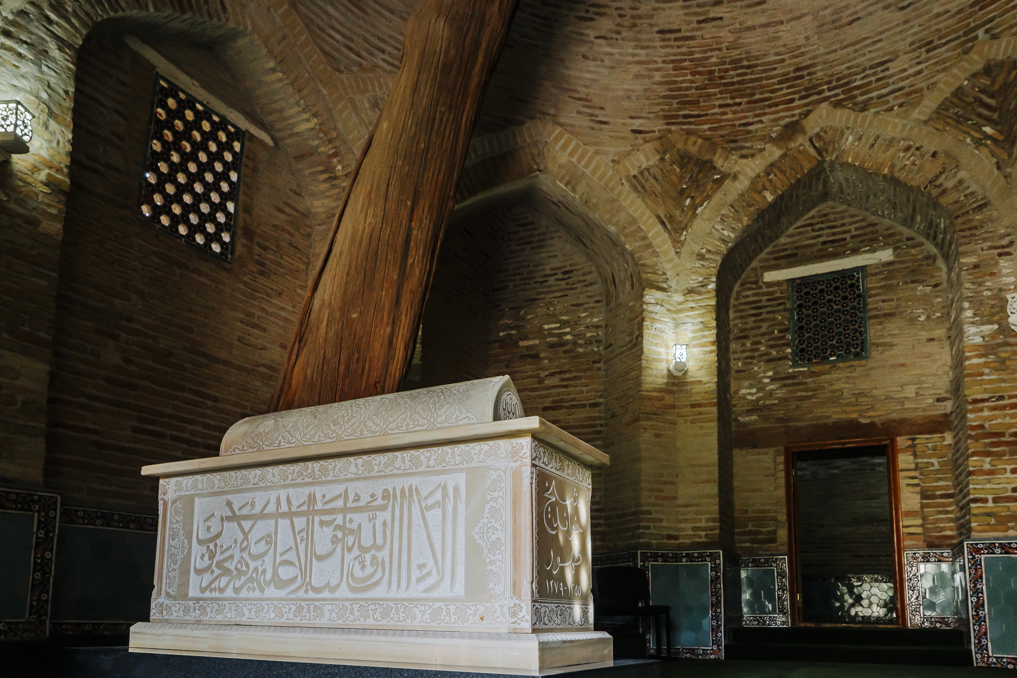 Intricate stone tomb with Arabic inscriptions inside a brick chamber featuring arched ceilings and wooden beams.