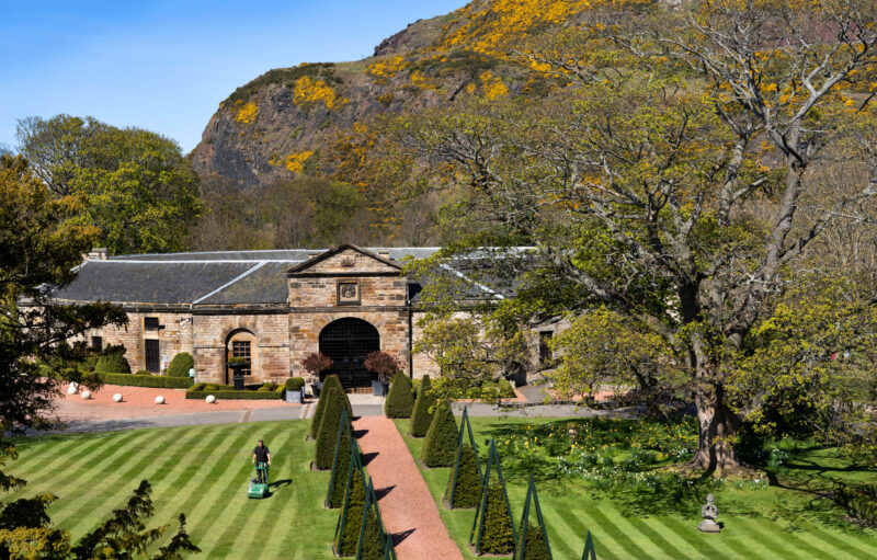 Historic stables building at Prestonfield House, surrounded by manicured lawns and trees, with a mountain backdrop.