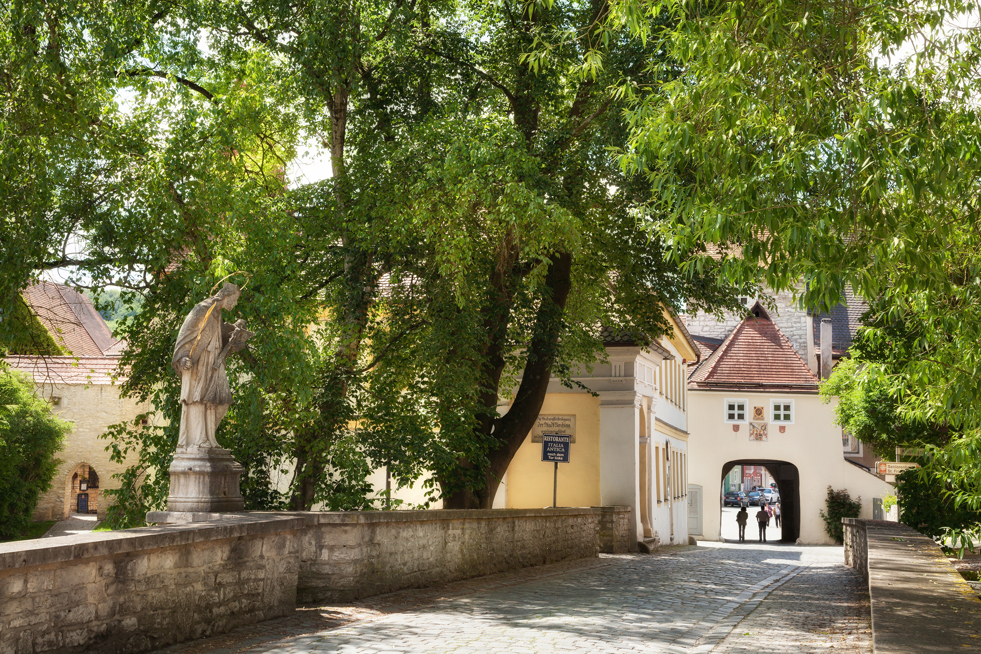 Statue of an angel beside a stone bridge leading to a white building with an archway, surrounded by lush green trees.