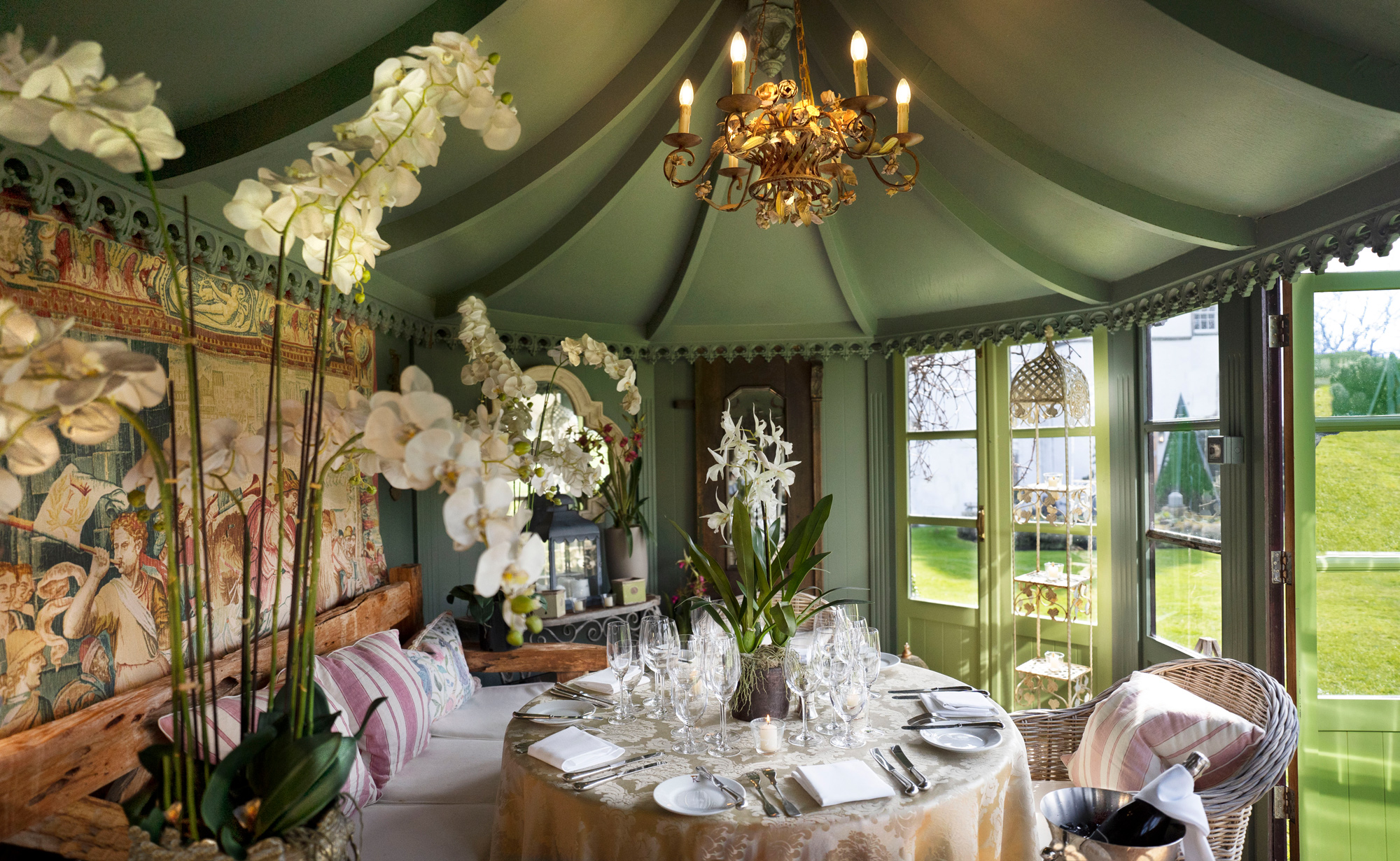 Elegant tea house interior featuring a round table set with glassware, surrounded by lush orchids and a chandelier.