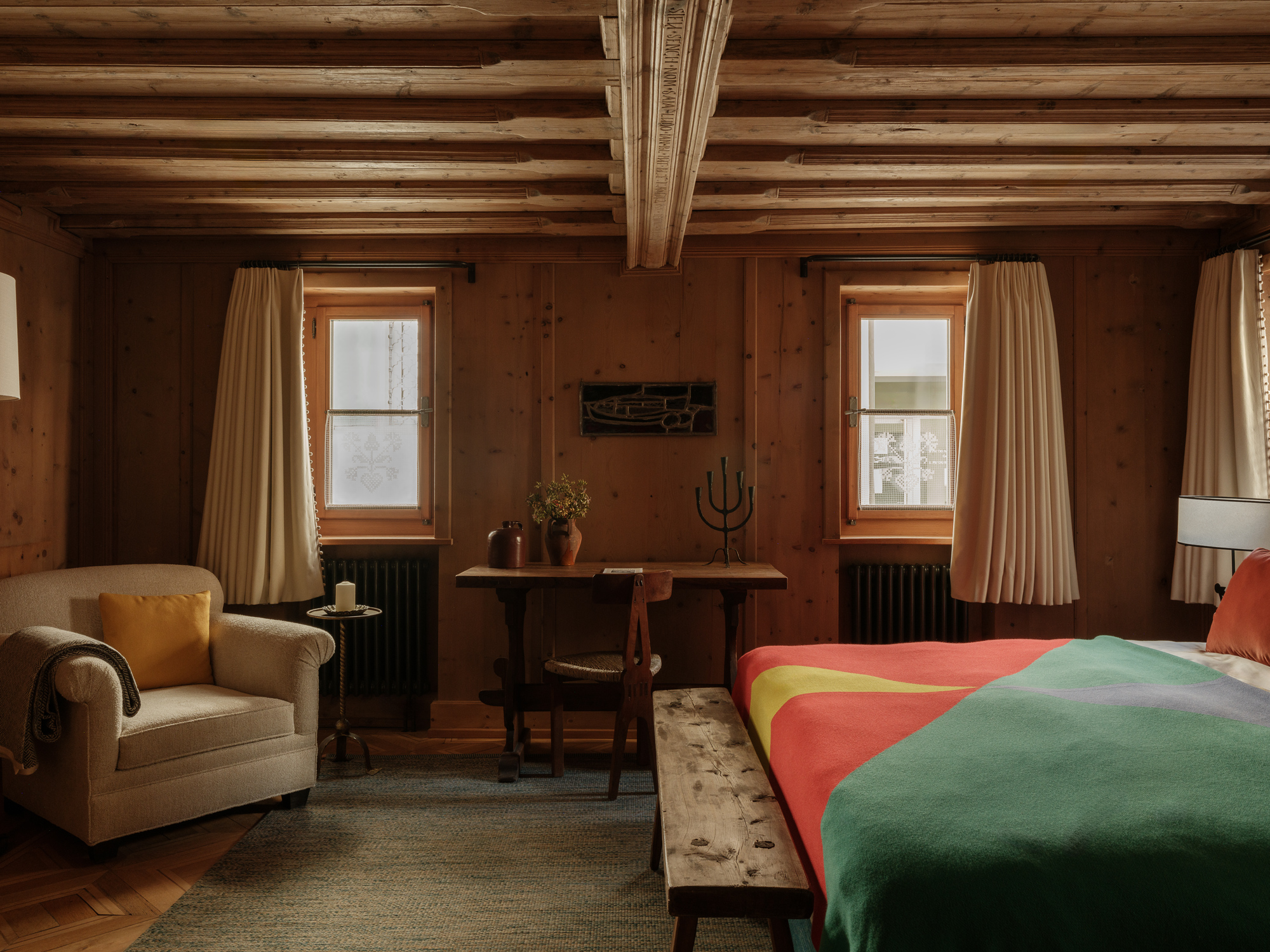 Cozy bedroom with wooden walls, colorful bedding, a sofa, and a small table by the windows.