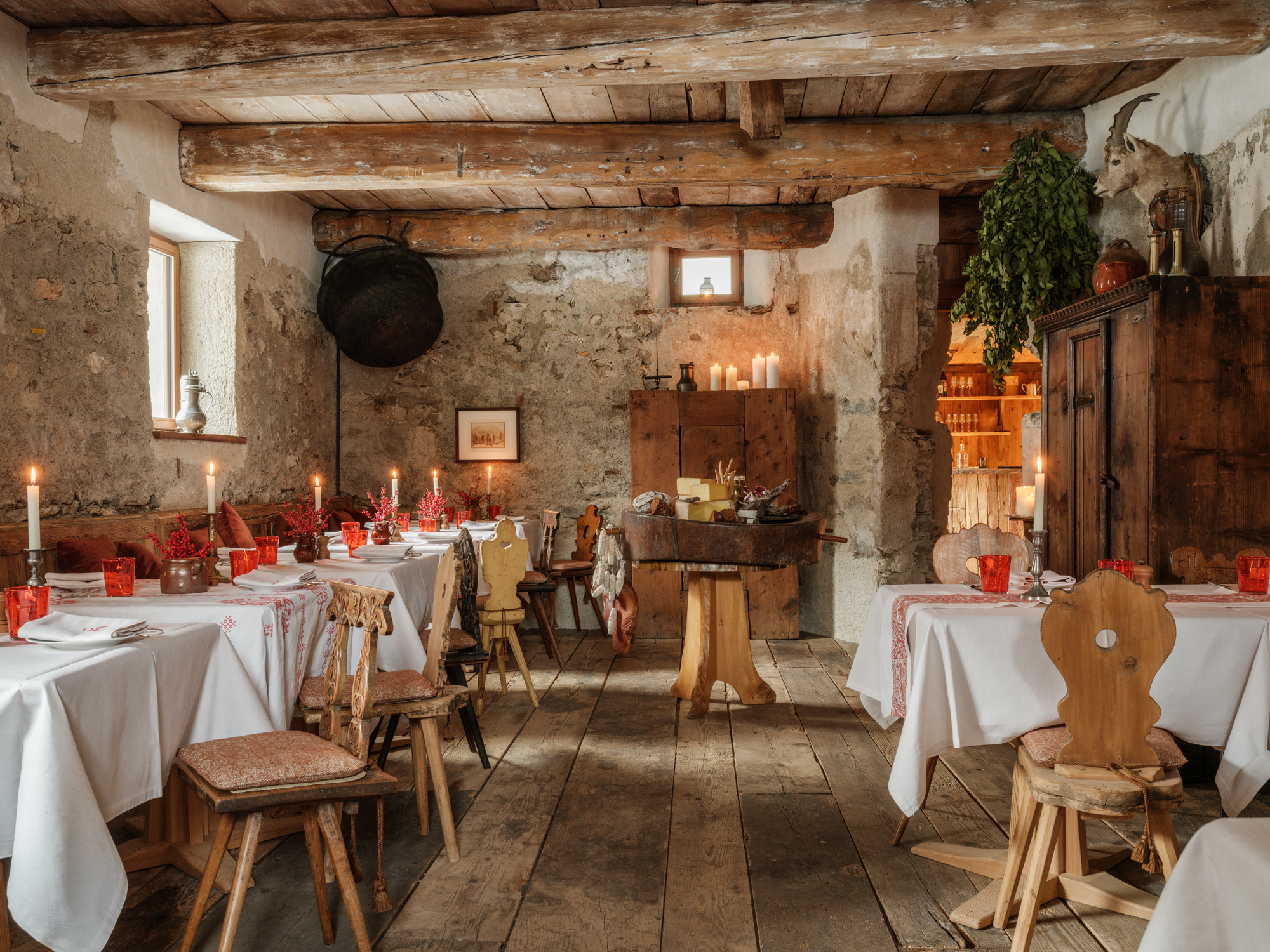 Rustic restaurant interior with wooden beams, tables set with white tablecloths, red glasses, and candles, creating a cozy...