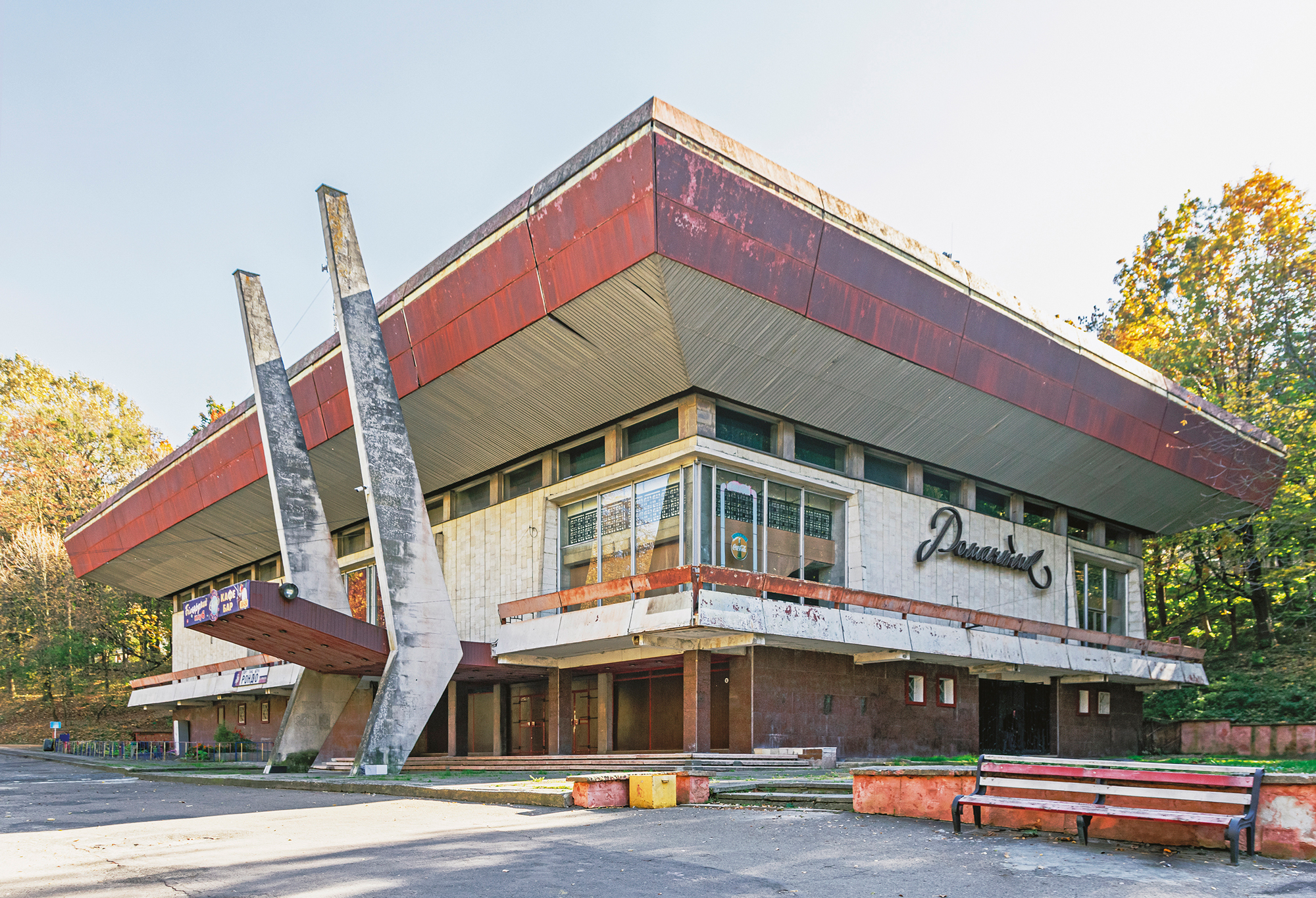 Abandoned modernist building with a flat roof and prominent support beams, surrounded by trees in autumn.