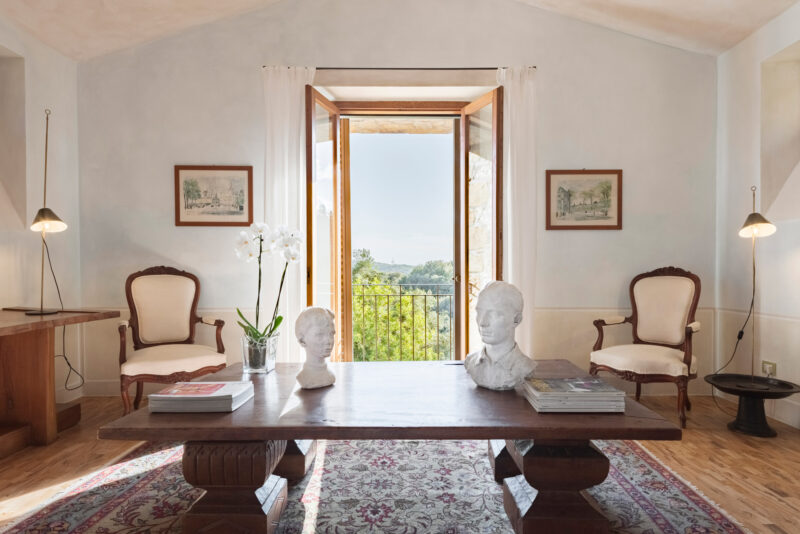 Elegant interior of Castello di Vicarello featuring a wooden table, two chairs, and sculptures, with a view of greenery ou...