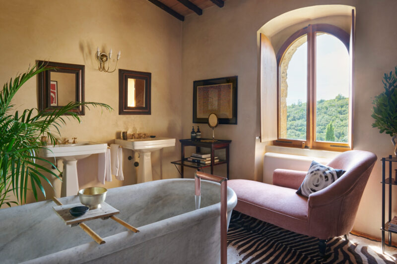 Elegant bathroom with a freestanding marble tub, pink armchair, and large window overlooking greenery, featuring decorativ...