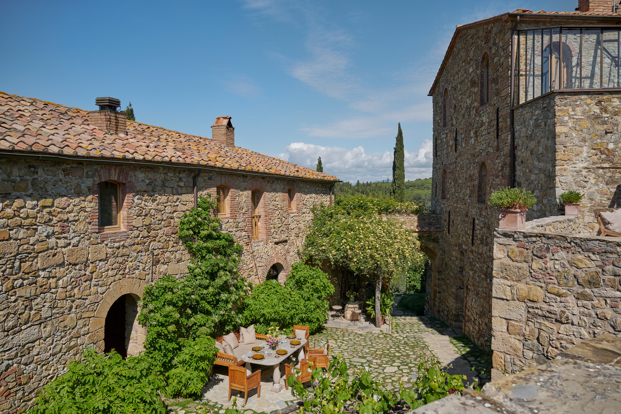 Rustic stone buildings surround a courtyard with a dining table set among lush greenery at Castello di Vicarello.