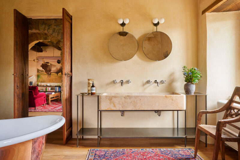 Elegant bathroom featuring a stone sink with two mirrors, vintage lighting, and a view into a cozy room with a red sofa.