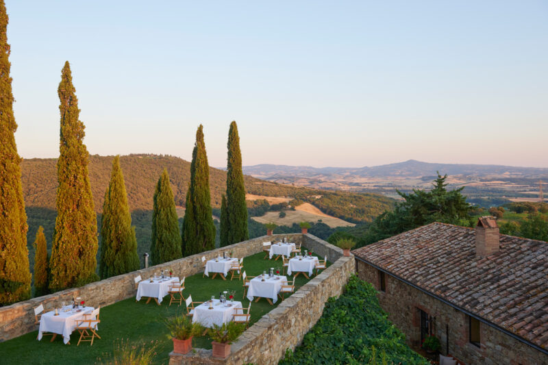 Outdoor dining setup with white tables on a terrace, overlooking rolling hills at sunset near Castello di Vicarello.