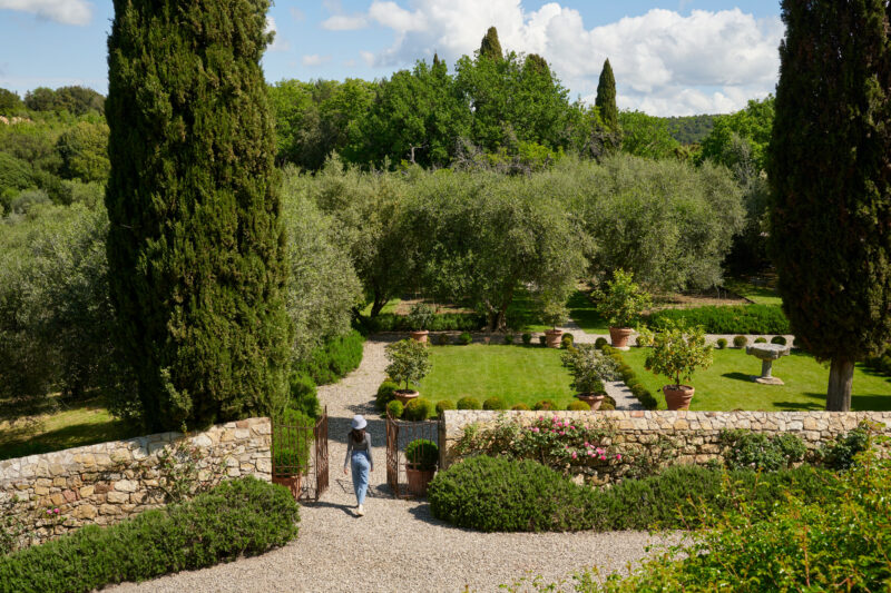 A woman in a wide-brimmed hat walks through a garden with olive trees and stone pathways at Castello di Vicarello.