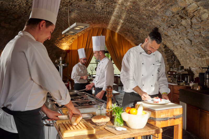 Chefs preparing food in a rustic kitchen with stone walls, featuring fresh ingredients and cooking equipment.