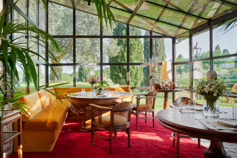 Brightly lit dining area with glass walls, featuring wooden tables, chairs, and a vibrant red carpet, surrounded by greenery.