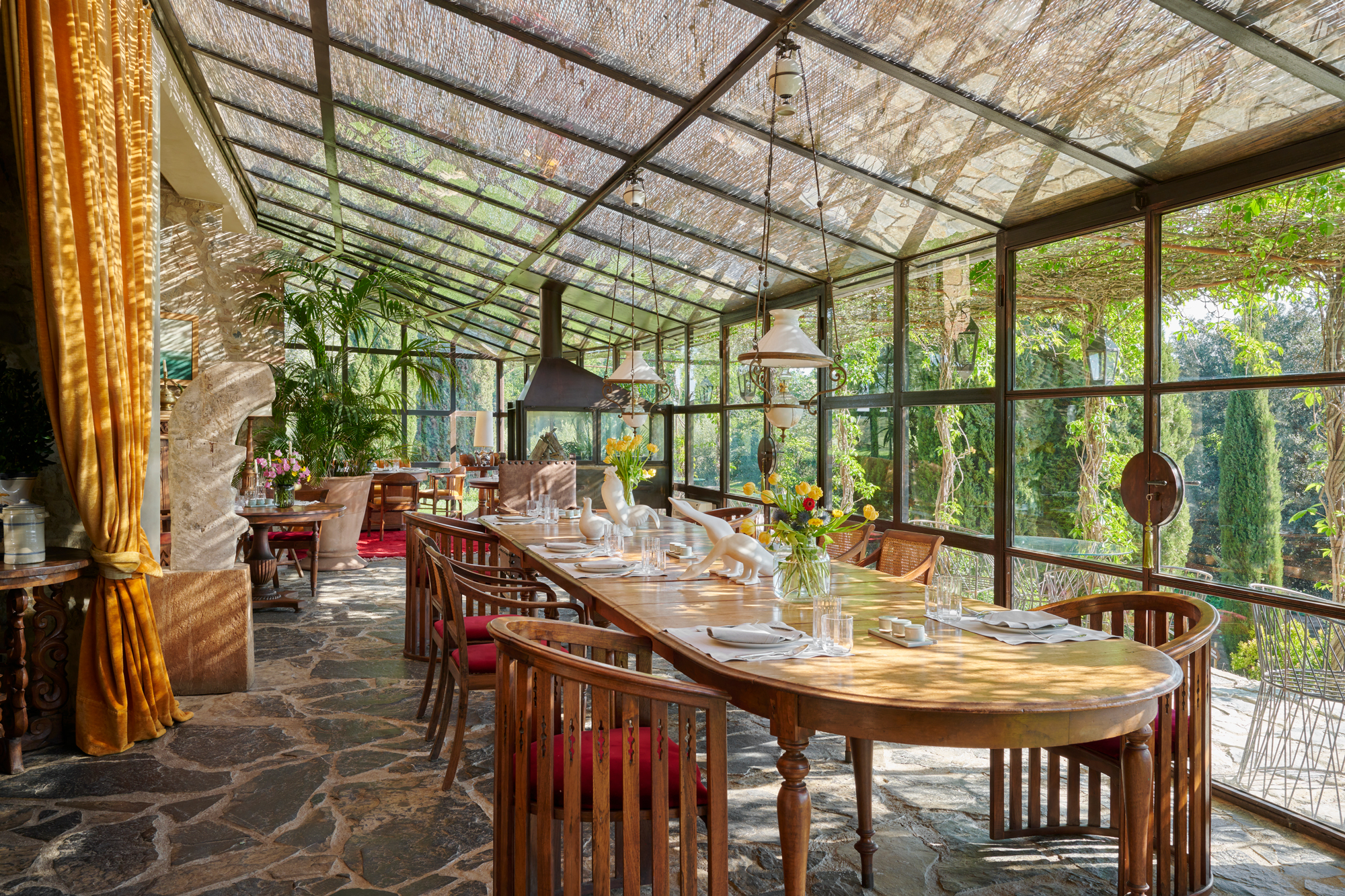 Bright, airy dining area with a wooden table set for a meal, surrounded by greenery and large glass windows.