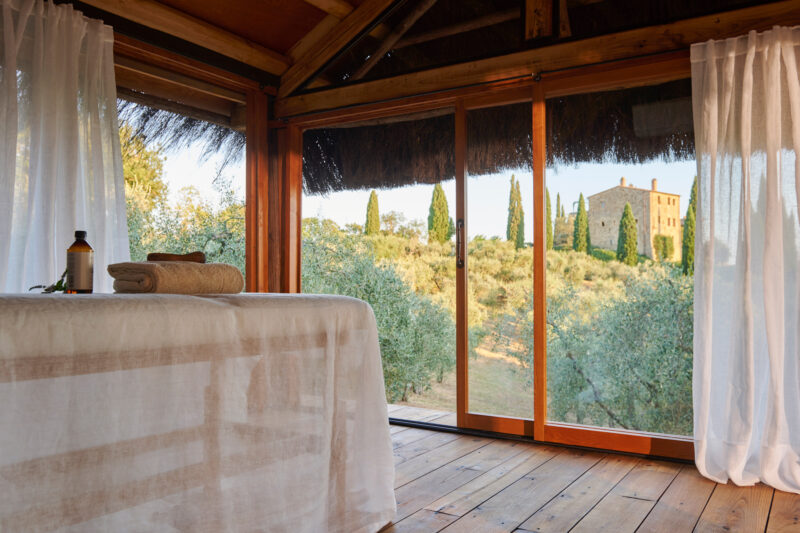 Spa room with a massage table and towels, overlooking olive trees and Castello di Vicarello in the background.
