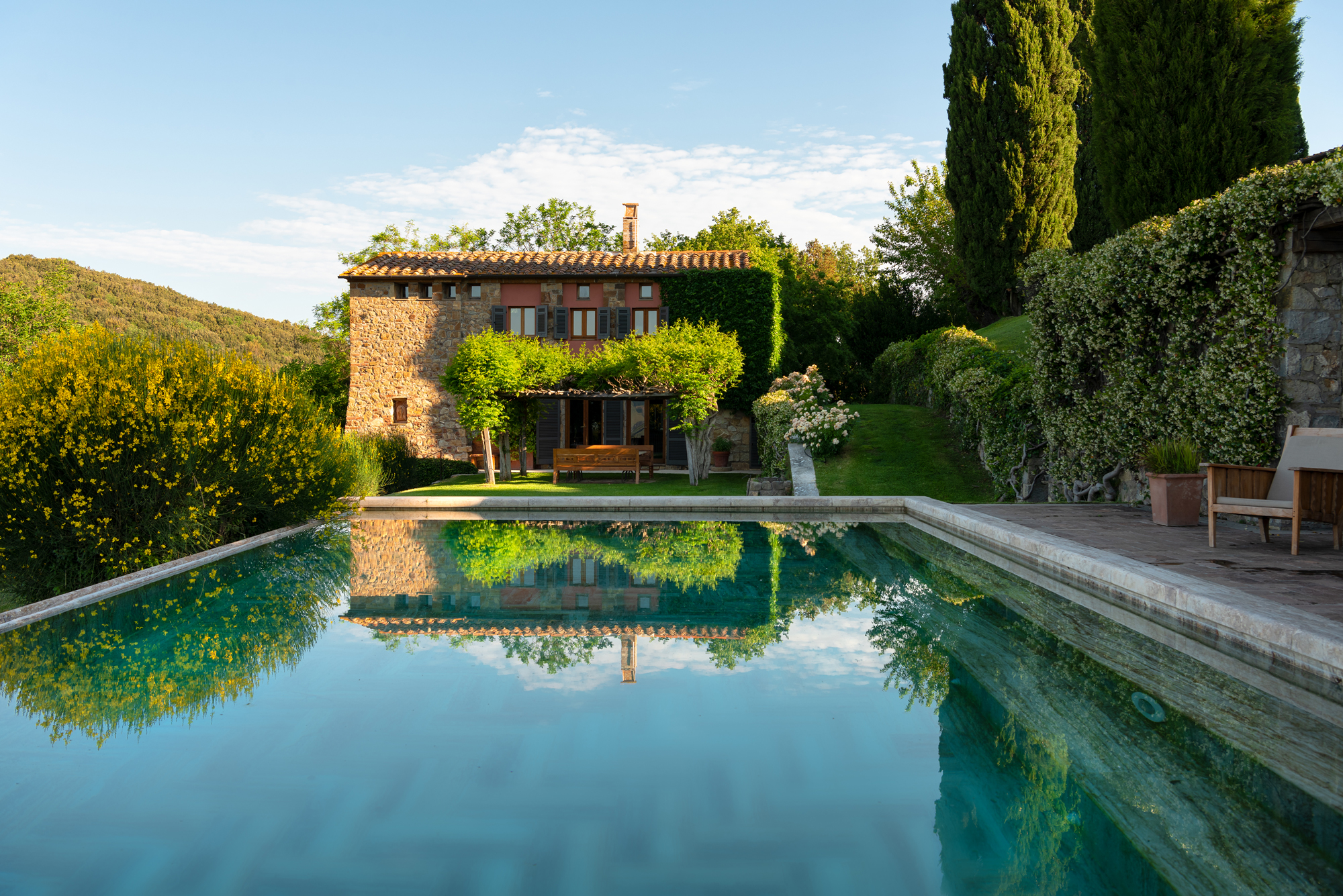Stone villa with lush greenery and a swimming pool reflecting the sky, surrounded by hills at Castello di Vicarello.