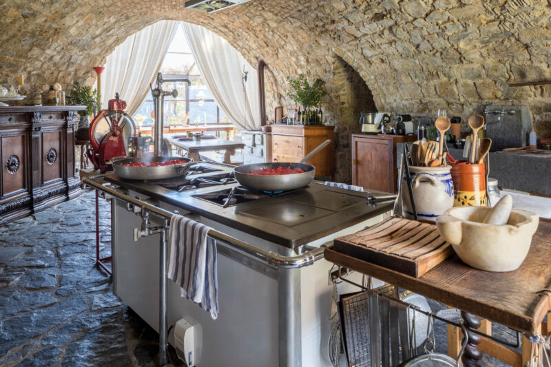 Rustic kitchen in Castello di Vicarello with stone walls, large stove, and wooden furniture, featuring cooking utensils an...