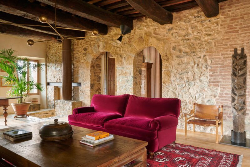 Cozy living room in Castello di Vicarello featuring a red velvet sofa, wooden beams, stone walls, and decorative plants.