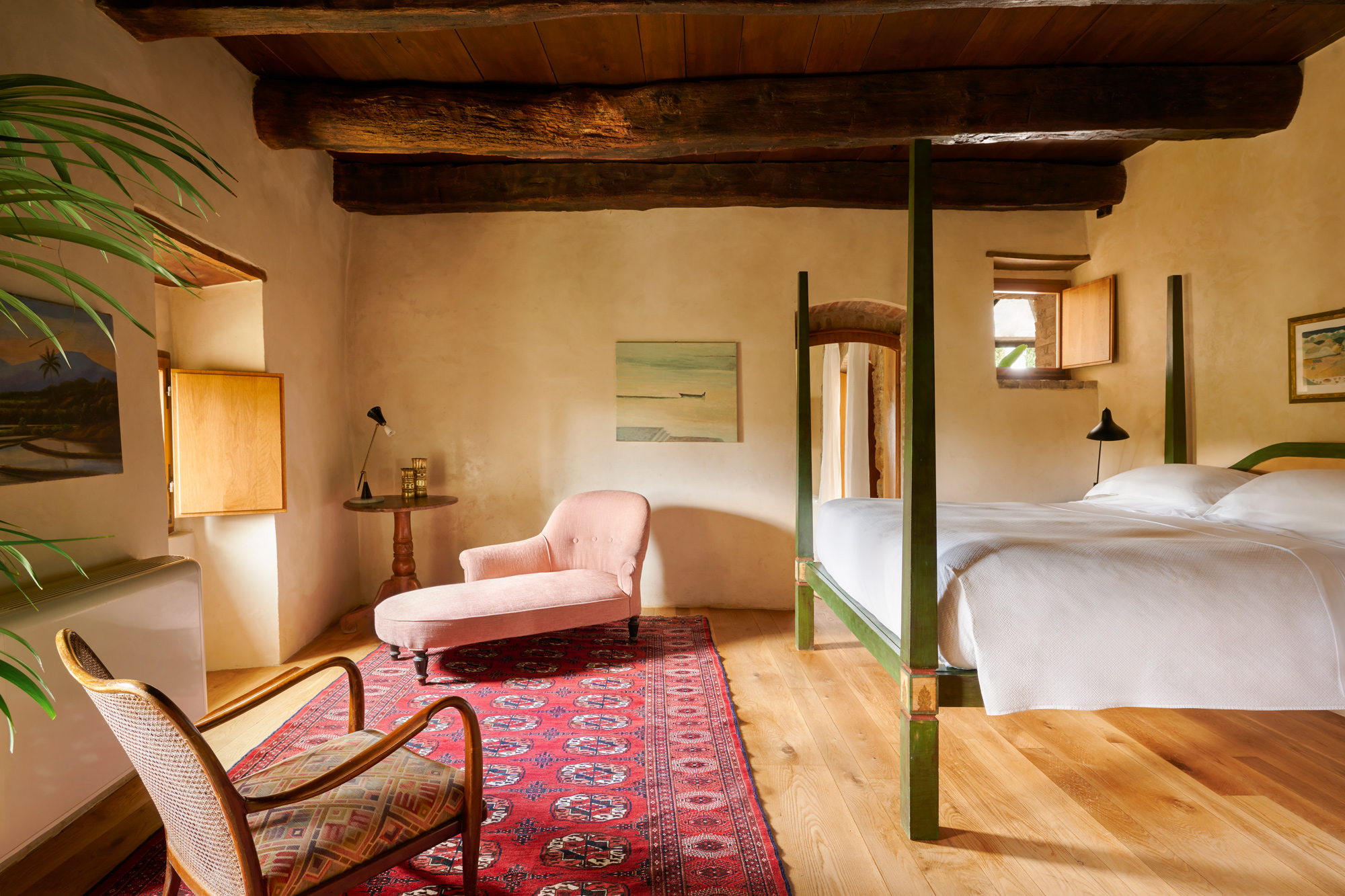 Cozy bedroom with a four-poster bed, pink armchair, wooden beams, and artwork, featuring warm tones and a rustic feel.
