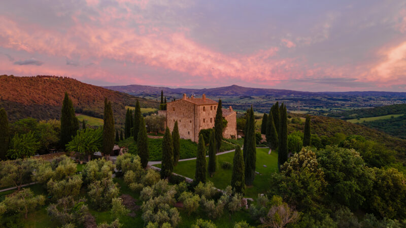 Castello di Vicarello at sunset, surrounded by cypress trees and olive groves, with rolling hills in the background.